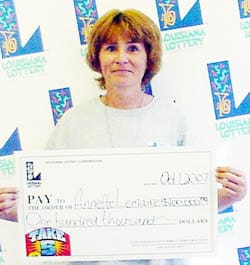 Woman holds an oversized check in front of a white background patterned with the Louisiana Lottery logo