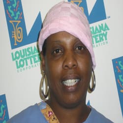 Woman with hoop earrings and a pink hat stands in front of a white background patterned with the Louisiana Lottery logo