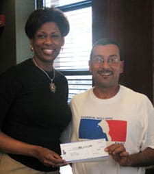 Man and woman hold a small check and smile for the camera in front of a bright window
