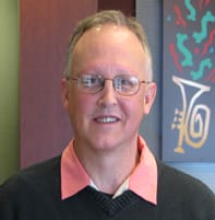 Man with a black sweater, pink collar, and glasses smiles in a Louisiana Lottery office