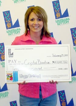 Woman with shoulder length hair and a pink shirt smiles with an oversized check in front of a white background patterned with the Louisiana Lottery logo