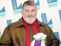 Man in a brown leather coat and red shirt smiles with a Louisiana Lottery prize bag in front of a white background patterned with the Louisiana Lottery logo