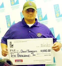 Man in a purple polo shirt and a green cap holds an oversized check in front of a faded white background patterned with the Louisiana Lottery logo