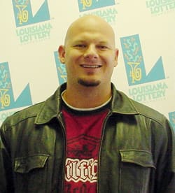 Bald man with goatee smiles in front of a faded white background patterned with the Louisiana Lottery logo