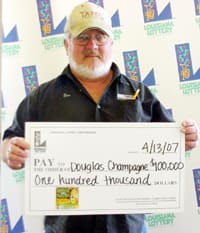 White-bearded man in black shirt and tan cap holds oversized check in front of a white background patterned with the Louisiana Lottery logo