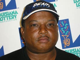 Man in black shirt and black Reno cap stands in front of white background patterned with the Louisiana Lottery logo
