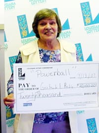 Older woman holds a giant check in front of a faded white background patterned with the Louisiana Lottery logo