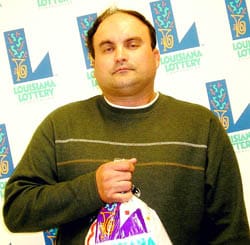 Man in a green sweater holds a Louisiana Lottery prize bag in front of a white background patterned with the Louisiana Lottery logo