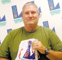 Man in a olive green tshirt smiles with a Louisiana Lottery prize bag in front of a white background patterned with the Louisiana Lottery logo