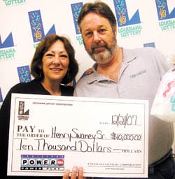 Man and woman hold an oversized check and smile in front of a white background patterned with the Louisiana Lottery logo