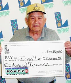 Older man in a gray collared shirt and yellow cap holds an oversized check in front of a white background patterned with the Louisiana Lottery logo