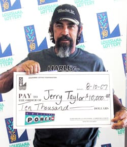 Bearded man in a black cap holds an oversized check in front of a white background patterned with the Louisiana Lottery logo