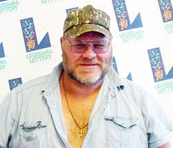 Man in a blue short sleeved button down shirt and camo hat smiles in front of a white background patterned with the Louisiana Lottery logo