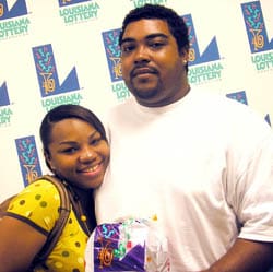 Man and woman hold Louisiana Lottery prize bag in front of white background patterned with the Louisiana Lottery logo