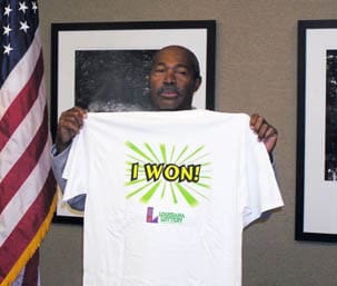 Man holds up a white tshirt reading "I Won!" in front of a plain wall with framed art and an American flag