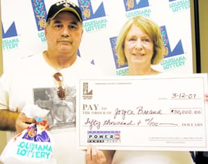 Man and woman hold oversized check and Louisiana Lottery prize bag in front of a white background patterned with the Louisiana Lottery logo