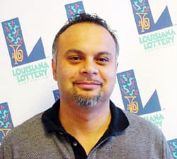 Man in a gray and black collared shirt stands in front of a white background patterned with the Louisiana Lottery logo