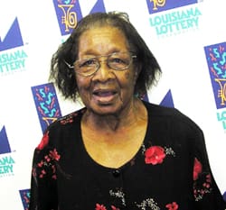 Older woman in a black floral shirt stands in front of a white background patterned with the Louisiana Lottery logo