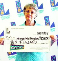 Woman in a blue shirt smiles with an oversized check in front of a white background patterned with the Louisiana Lottery logo