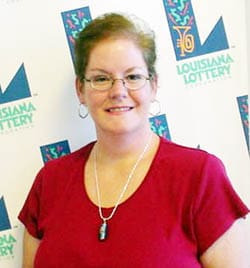 Short-haired woman in a red shirt and glasses smiles in front of a white background patterned with the Louisiana Lottery logo