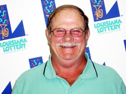 Mustached man in an aqua polo smiles in front of a white background patterned with the Louisiana Lottery logo