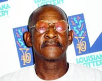 Man with red sunglasses stands in front of a white background patterned with the Louisiana Lottery logo
