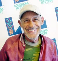 Man in a red silk members only jacket and camo tshirt smiles in front of a white background patterned with the Louisiana Lottery logo