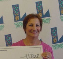 Woman with short hair and a pink shirt smiles in front of a white background patterned with the Louisiana Lottery logo