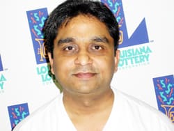 Man in a white v-neck tshirt stands in front of a white background patterned with the Louisiana Lottery logo