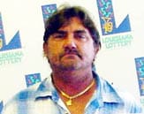 Man in a blue plaid shirt and gold chain stands in front of a white background patterned with the Louisiana Lottery logo