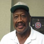 Man in a white collared shirt and a cap smiles for the camera in front of a light green wall