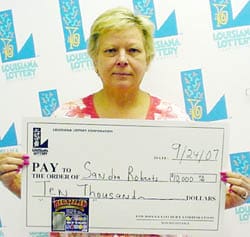 Woman with short light hair holds an oversized check in front of a faded white background patterned with the Louisiana Lottery logo