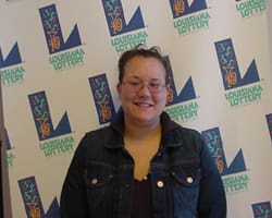 Woman in a black jacket and top smiles in front of a white background patterned with the Louisiana Lottery logo