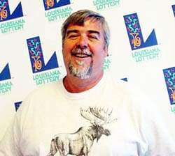 Man in a white tshirt with a moose on it laughs in front of a white background patterned with the Louisiana Lottery logo