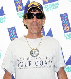 Man in a white tshirt and purple LSU hat smiles in front of a white background patterned with the Louisiana Lottery logo