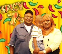 Man, woman, and young boy pose in front of an orange digital background with red and green cartoon chili peppers and "Spicy 7" written in flames