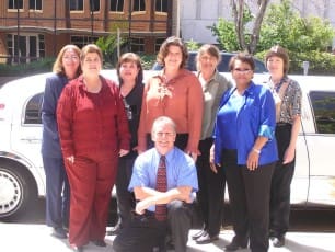 Group of men and women pose outdoors on the sidewalk in front of a white stretch limosuine