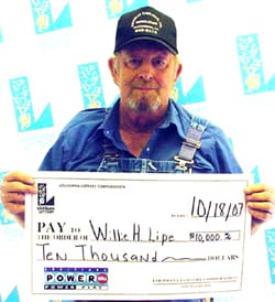 Man in overalls and a black cap holds an oversized check in front of a faded white background patterned with the Louisiana Lottery logo