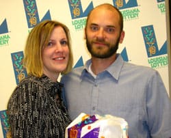 Man and woman smile with Louisiana Lottery prize bag in front of white background patterned with the Louisiana Lottery logo